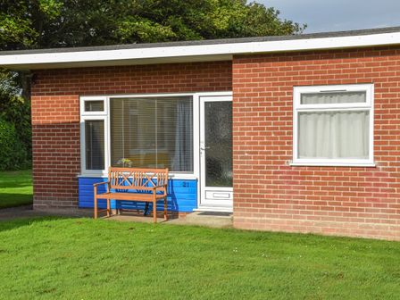 A brick house with a white door two windows and a wooden bench on the grass at 21 Siesta Mar Mundesley