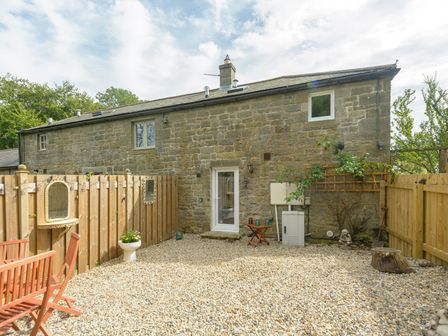 A stone house with a small fenced gravel yard and outdoor wooden chairs and table at Rose Cottage in Edlingham