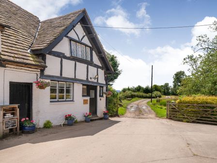 A white cottage with black timber framing and a tiled roof next to a dirt road with a wooden gate and green bushes at Fern Hall Cottage Whitney-on-Wye near Hay-on-Wye