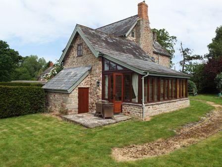 An exterior view of a stone house with a conservatory and a small patio with chairs on a grassy lawn at The Granary in Walford near Ross-on-Wye