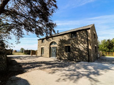 A stone house with a grey door and windows under a blue sky with trees and a wooden gate at Damson Barn in Kirkby Lonsdale