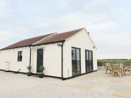 A single story house with white walls and a tiled roof next to a paved patio with wooden outdoor furniture at Fieldside in Hunmanby