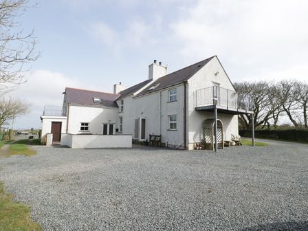 A two-story white house with a gravel driveway and a balcony surrounded by trees at Bodegri Cottage Llanrhyddlad near Church Bay