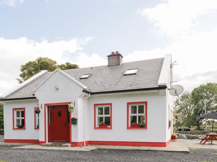A white cottage with red trim and a red door with a satellite dish and outdoor seating area at Lough Mask Road Fishing Cottage in Ballinrobe County Mayo