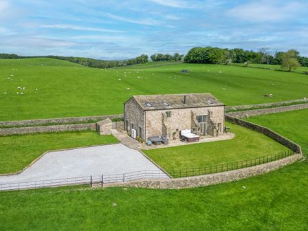 A stone house with a patio and hot tub surrounded by green fields and stone walls at Crane Field Laithe in Otterburn North Yorkshire