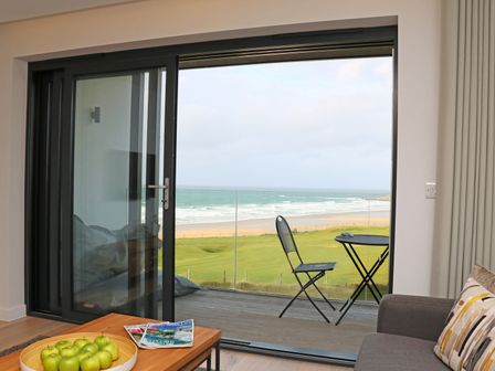 A living room with a bowl of green apples on a table and a sliding glass door opening to a balcony with a chair and table overlooking the ocean at Little Fistral in Fistral