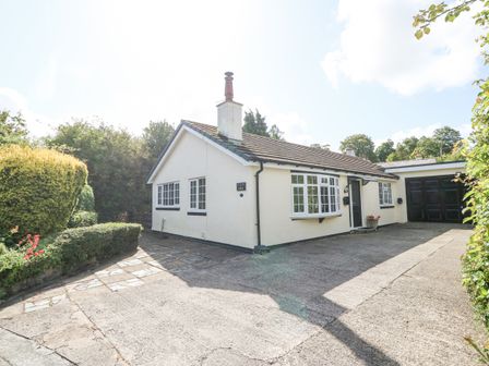 A white single-story house with black trim windows a driveway a garage and surrounding greenery at Glan Dwr in Red Wharf Bay
