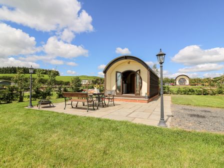 A small curved cabin with double doors open on a paved patio with a metal table and bench and two street lamps in a green field at Meadow View in Rhayader