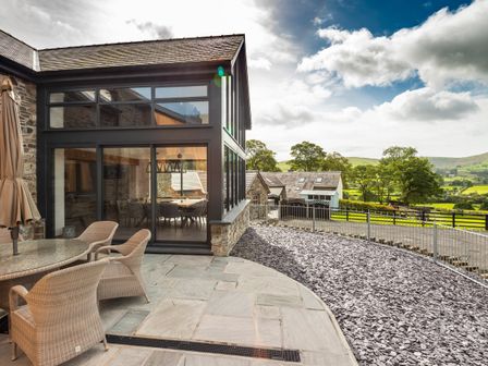 An outdoor patio area with wicker chairs and a glass table next to a stone building with large windows and a rural landscape at Granar in Llangwm near Bala