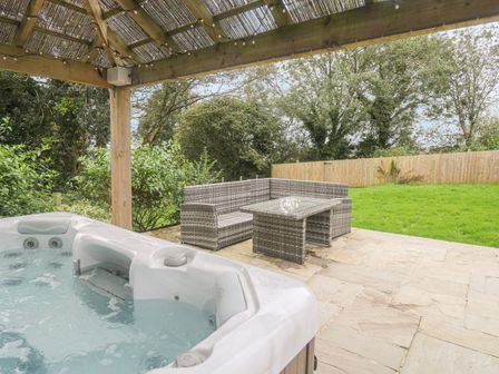 An outdoor patio with a hot tub under a wooden pergola and wicker seating with a table on a stone floor and grass and trees in the background at Kelyow in St Day