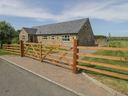 A single-story stone cottage with a gray roof behind a wooden gate and fence on a paved driveway at Rede River Cottage in West Woodburn