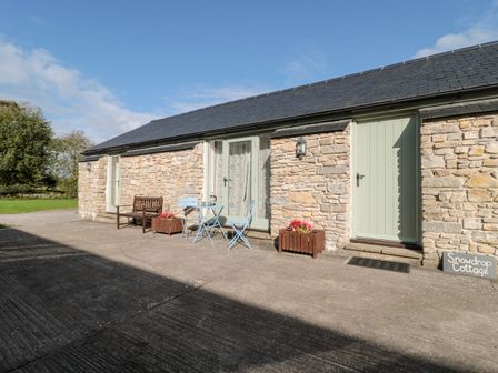 An exterior view of a stone cottage with two light green doors a small blue metal table and chairs and a wooden bench with flower planters at Snowdrop Cottage in Llantwit Major
