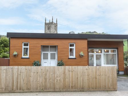 A wooden house with white doors and windows behind a wooden fence with a church tower in the background at The Cabin in Combe Martin