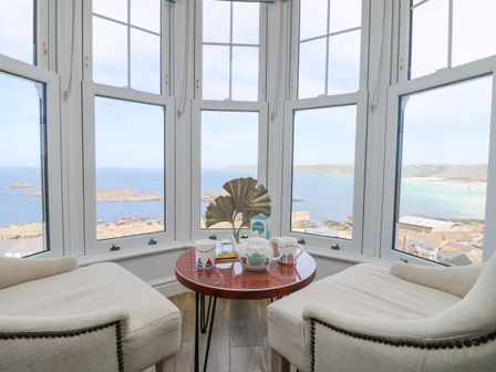 A seating area with two beige chairs and a round wooden table by large bay windows overlooking the sea at Sennen Heights in Sennen Cove near Penzance