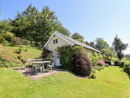 A small house with outdoor table and chairs on a patio surrounded by trees and bushes at Dolgenau Hir The Barn in Llawr y Glyn near Trefeglwys