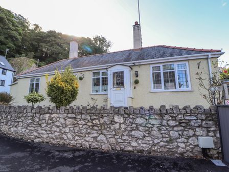 A single-story house with a tiled roof and two chimneys behind a stone wall with shrubs at Llidiart Cerrig in Dyserth