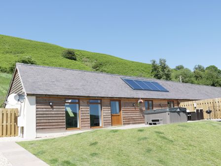 A single-story building with wooden siding windows and a door with a hot tub and outdoor seating on a grassy area at 1 Beacon View Barn in Beguildy near Knighton