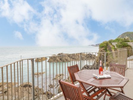 A balcony with a wooden table and chairs overlooking rocky coastline and sea at Oceans Side in Ilfracombe