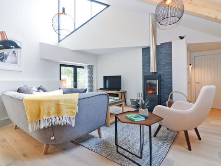 A living room with a grey sofa and beige armchair next to a wood burning stove at Long House Boskensoe Barns in Mawnan Smith