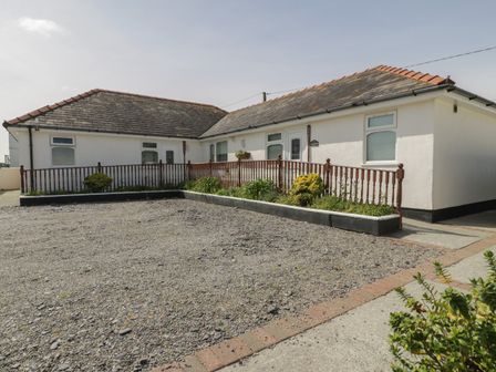 The exterior of a single-story house with white walls and a tiled roof with a gravel driveway and a wooden railing with plants along the front at Sea Spray in Dinas Dinlle