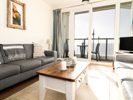 A living room with grey sofas a wooden coffee table and glass doors opening to a balcony with a sea view at 12 West End Point in Pwllheli