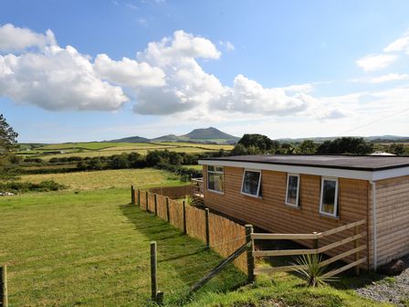 A wooden cabin next to a fenced grassy field with hills and clouds in the background at Y Caban Clyd in Morfa Nefyn