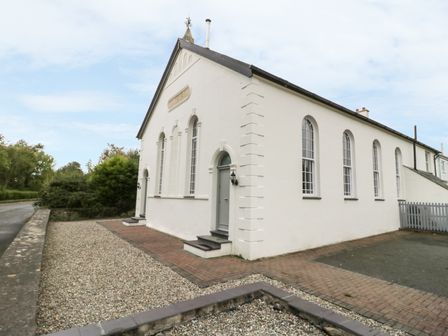 The exterior of a white building with arched windows and two doors with steps surrounded by a gravel and paved area at Capel Bryn Bachau in Chwilog near Criccieth
