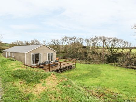 A single-story building with beige siding and a wooden deck on a grassy area with trees in the background at Nant Y Felin Lodge in Aberdaron