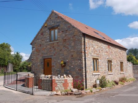 A stone building with a tiled roof and wooden door and windows surrounded by a low stone wall at The Old Cider Barn in Wookey