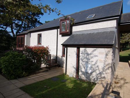 An exterior view of a two-story house with a slate roof a bench outside surrounded by bushes at Pine Cottage in Maenporth