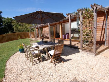An outdoor dining area with table chairs and umbrella on a gravel patio next to a wooden house at Orchard Retreat in Sidmouth