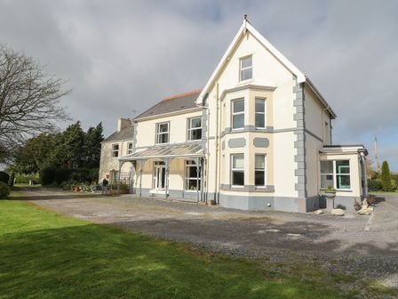 An exterior view of a large two-story house with a covered porch and a bay window with a gravel driveway and lawn at Muriau Park in Caernarfon