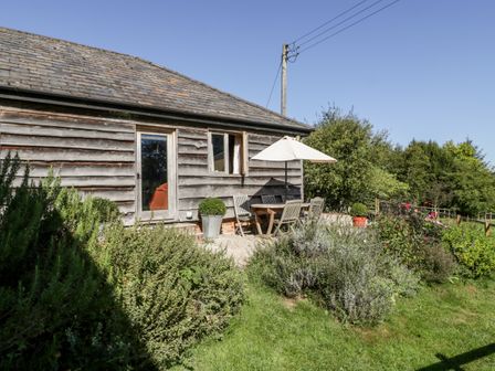 An outdoor patio with wooden furniture and a white umbrella next to a wooden building surrounded by bushes at The Old Cart Shed in Rockbourne near Fordingbridge
