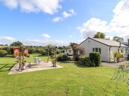 A garden with a white patio table and chairs under a wooden pergola and a small white house in the background at The Flat At Brwyn in Brynford