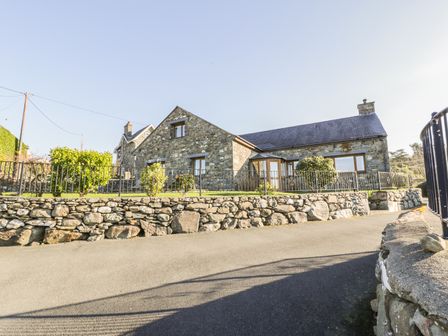 A stone house with a sloped roof and fenced garden beside a paved road at Awel Y Llan in Llanfair