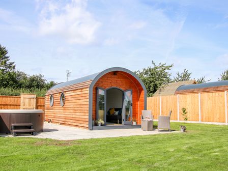 A wooden pod with open doors on a patio with chairs and a hot tub in a fenced garden at Withy Willows in Diddlebury near Ludlow