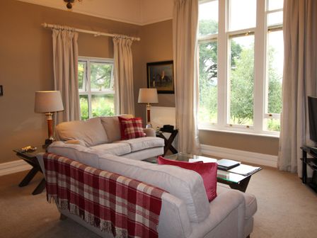 A living room with two beige sofas with red cushions and blankets two lamps a glass coffee table and large windows at Geltsdale Garden Apartment in Wetheral near Carlisle