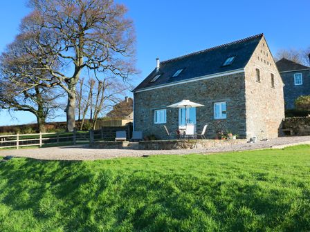 A stone house with outdoor furniture and umbrella on a patio near large trees and grass at Polly's Bower in Landrake