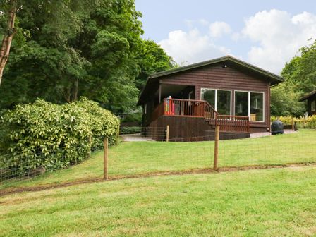 A wooden cabin with large windows on a grassy slope surrounded by trees and bushes at Kipling Lodge in Rudyard