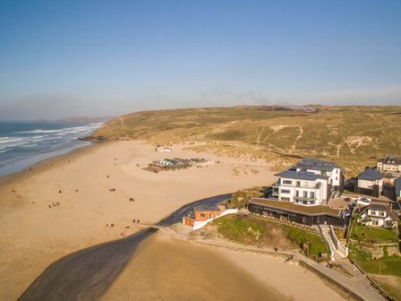 A sandy beach with scattered people a building complex on a cliff and hills in the background at Chapel Rock View The Dunes in Perranporth