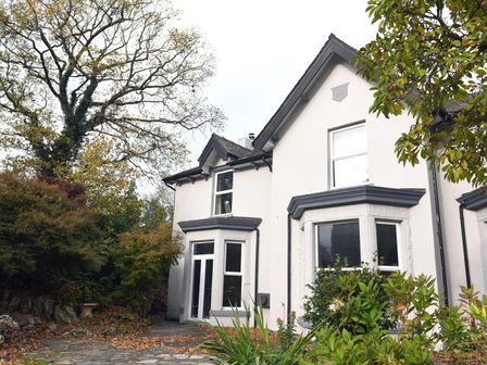 The exterior of a two-story white house with bay windows and surrounding trees and bushes at Ellie's Lodge in Ulverston