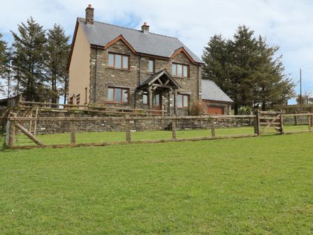 A two-story stone house with a wooden gate and fence on a grassy lawn with trees behind at Blaen Henllan in Builth Wells