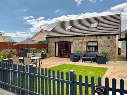 A stone cottage with a patio, outdoor table with umbrella and chairs, a lawn, and a black picket fence at Pottery Cottage Begelly near Kilgetty