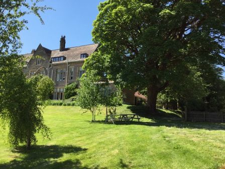 A large stone building with multiple windows behind trees and a grassy lawn with a picnic table at Geltsdale East Wing in Wetheral