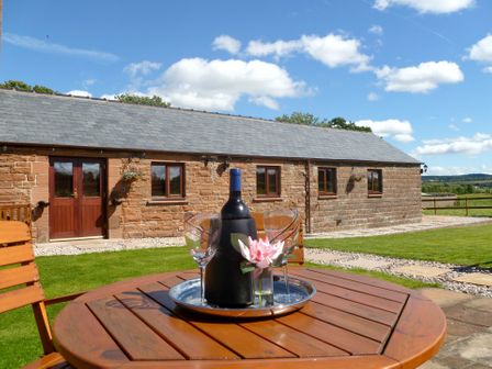 A wooden outdoor table with a bottle of wine and two glasses in front of a stone cottage under a blue sky at Daisy Cottage in Catterlen