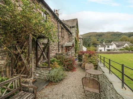A stone cottage exterior with a wooden bench metal table and chairs along a gravel path with greenery and a field beyond at Tanner Croft Cottage in Grasmere