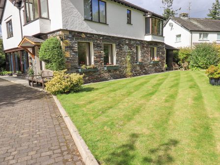 An exterior view of a house with stone walls and white upper walls a lawn with stripes and a paved driveway at Stonegarth Cottage in Grasmere