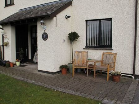 The front porch of a cottage with wooden chairs and a small table on a paved area with plants around at Thyme Cottage in Bowness-on-Windemere