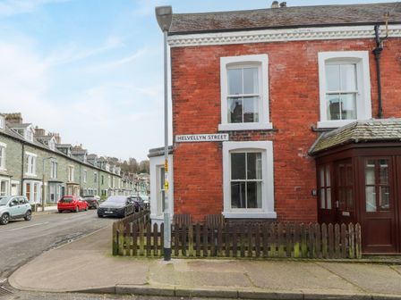 A red brick corner house with white framed windows and a small wooden fence on Helvellyn Street in Keswick