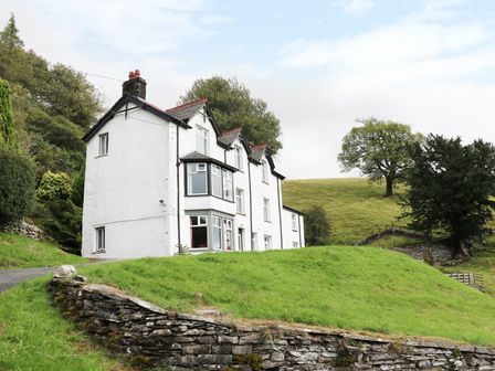 A white multi-story house with bay windows on a grassy hill surrounded by trees at Low Skelgill in Troutbeck